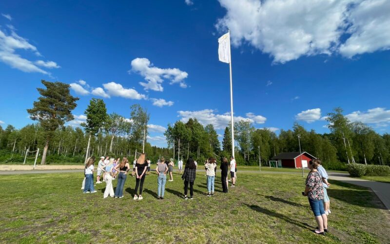 Nuoret nostavat Suomenlipun salkoon rippileirillä. Ryhmä nuoria kokoontuu lipputangon ympärille.