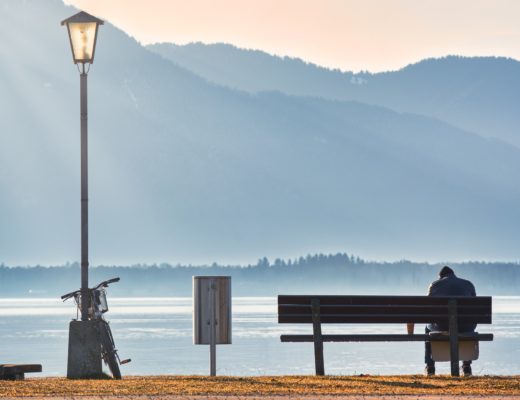 Ihminen istuu yksin penkillä järven rannalla. Taustalla näkyy sinisävyisiä vuoria.