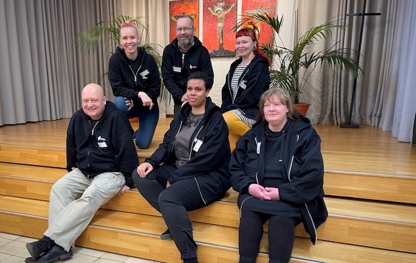 Kuusi hymyilevää Helsingin seurakuntayhtymän kokemusasiantuntijaa istuu ryhmäkuvassa portailla.
